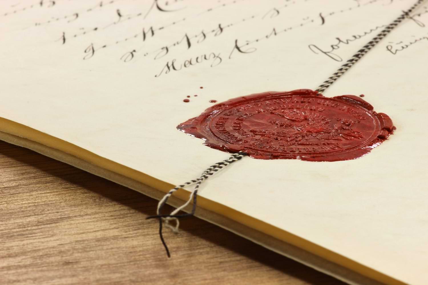 Ein altes Dokument mit kursiver Handschrift und einem roten Wachssiegel, das mit einer Kette gesichert ist.